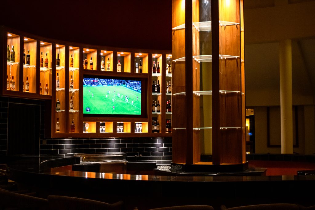 Bar counter at Anga Sky Bar, featuring warm wooden shelving, a well-stocked spirit display and a television screen above.