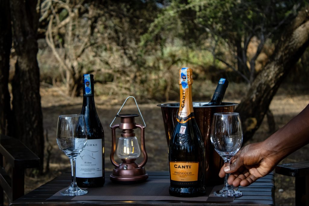 Wine, Prosecco, wine glasses, and a hurricane lantern set up for a luxury bush dining experience.