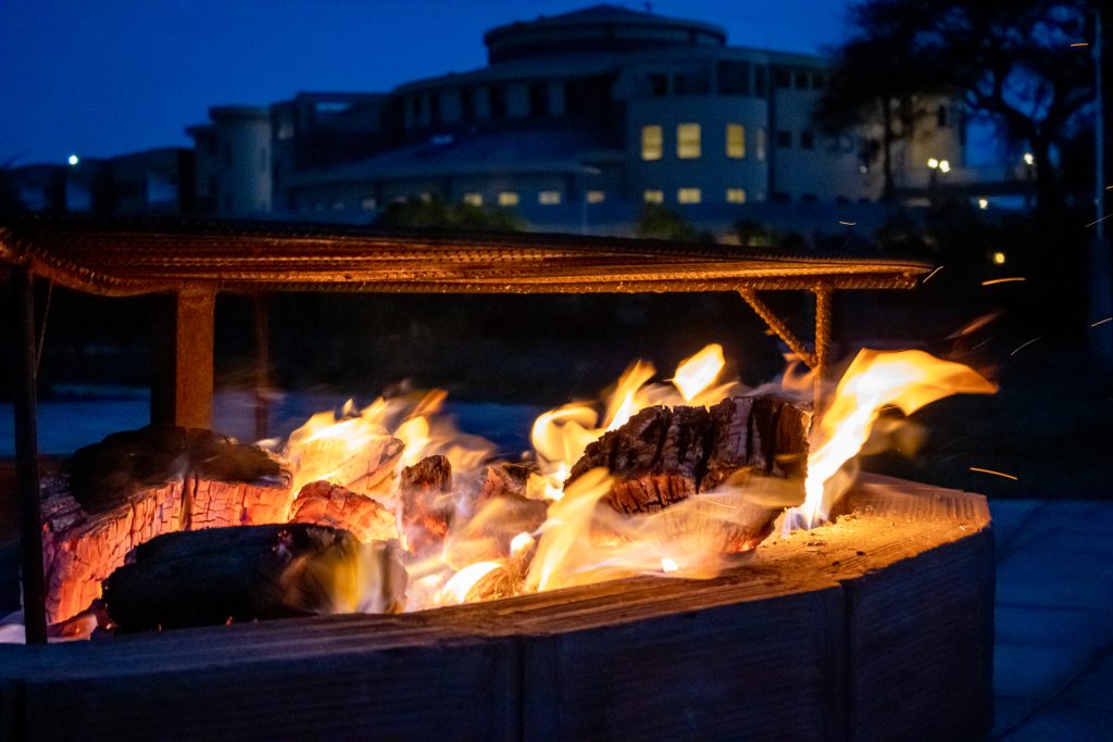 Enkima Firepit glowing with a wood fire at dusk, with the lodge illuminated in the background.