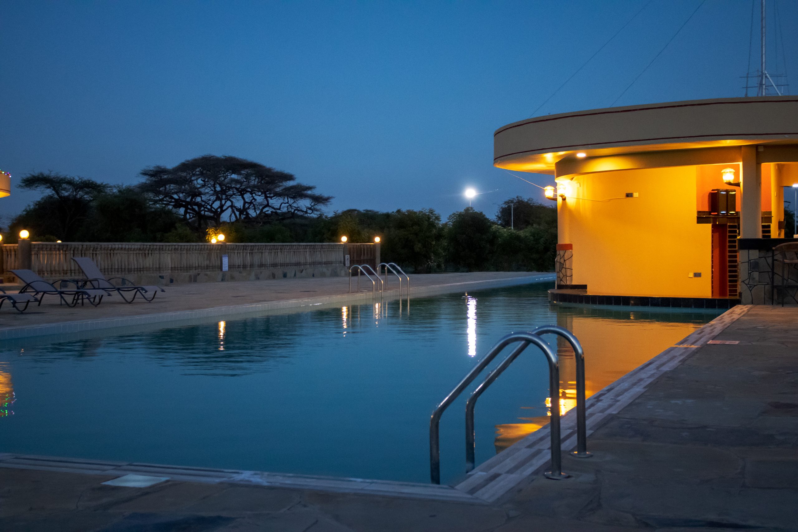 Illuminated Olkeju Pool Bar and swimming pool deck at dusk with acacia trees in the background.
