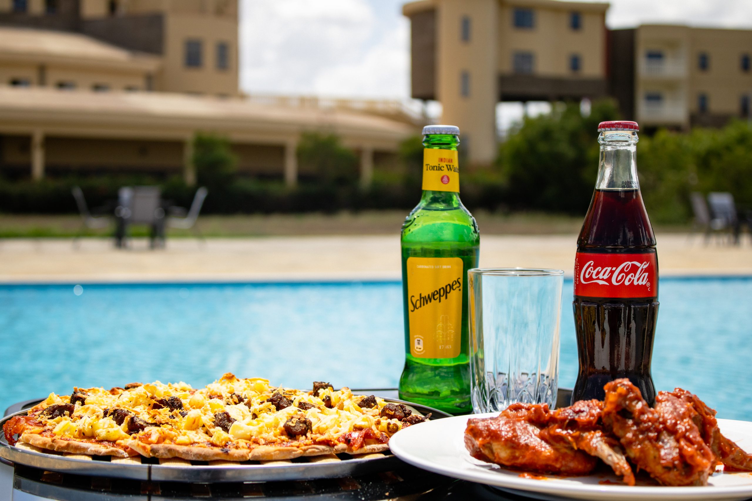 Close-up of poolside dining options including pizza, glazed chicken wings, and bottled soft drinks by the clear blue pool.